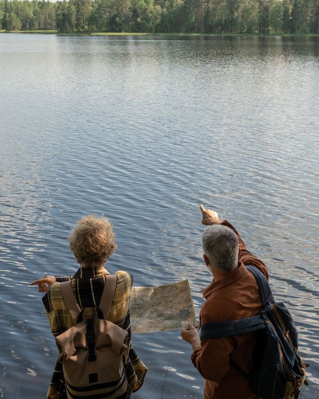 Casal de idosos com mochilas olhando um mapa à beira de um lago